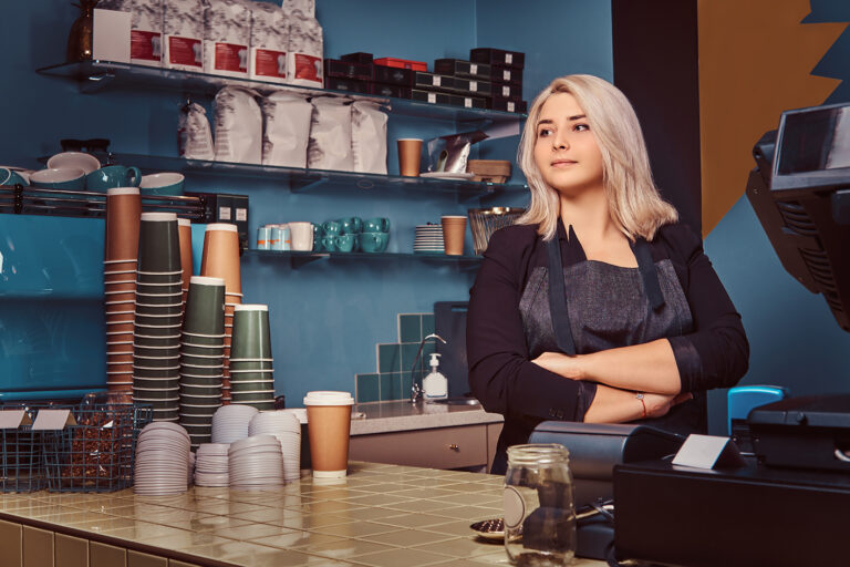 Confident female barista in apron standing with crossed arms at her UK coffee shop